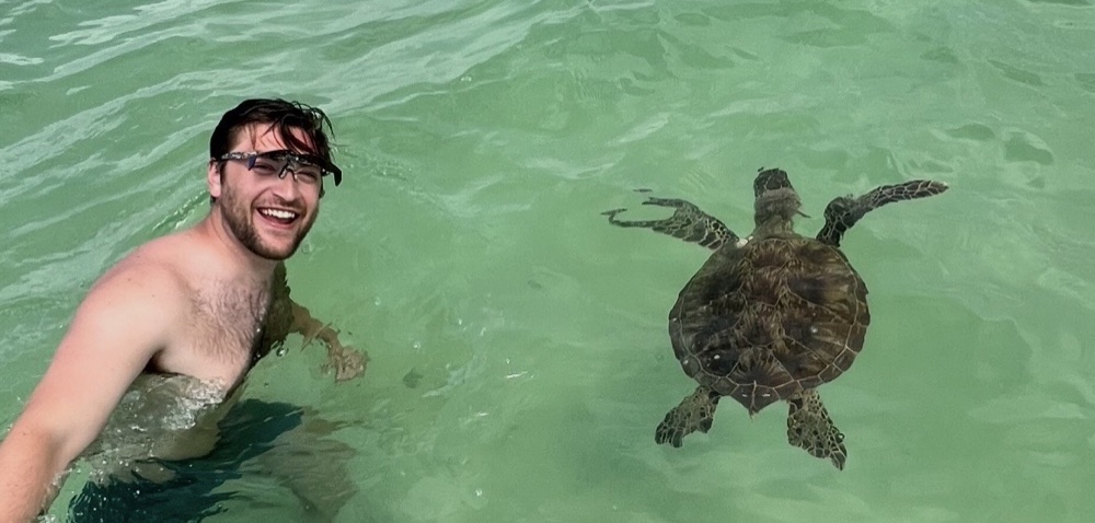James Palmer smiling in clear green water beside a swimming sea turtle